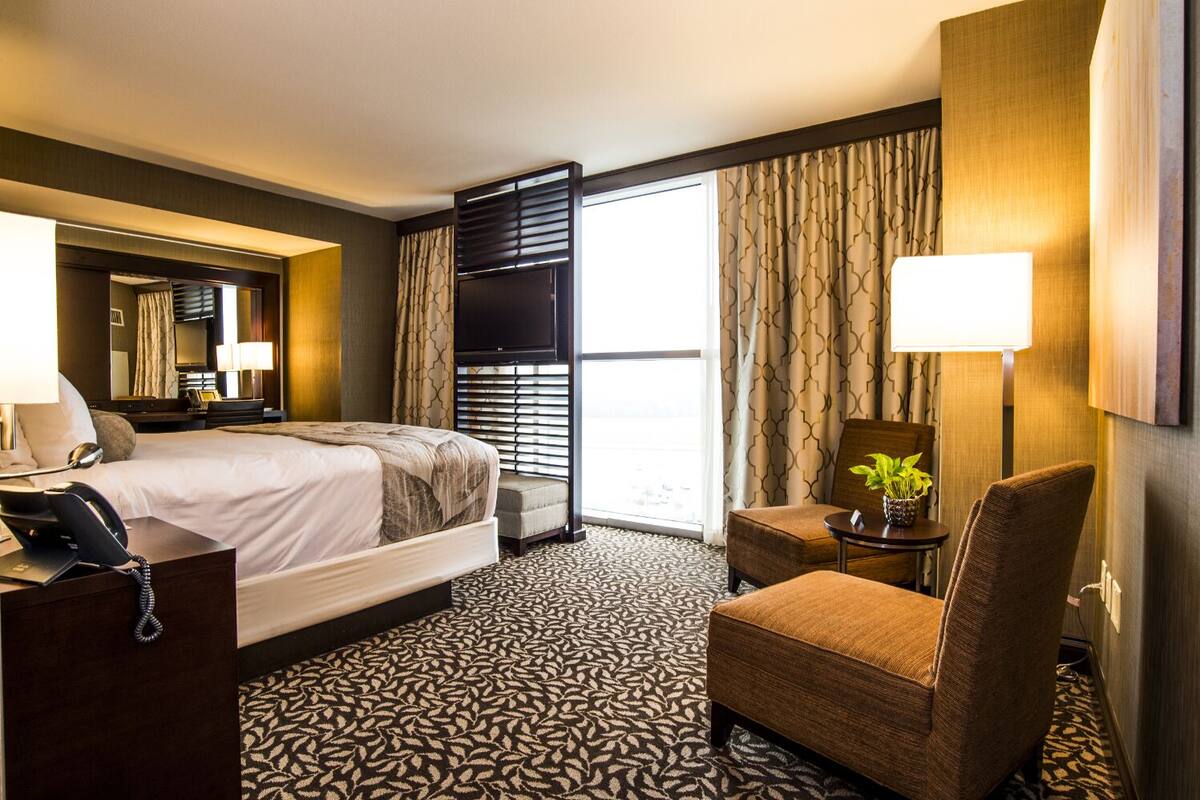 A hotel room with a bed, a desk with a telephone, a chair, a small table with a plant, and a large window with curtains.
