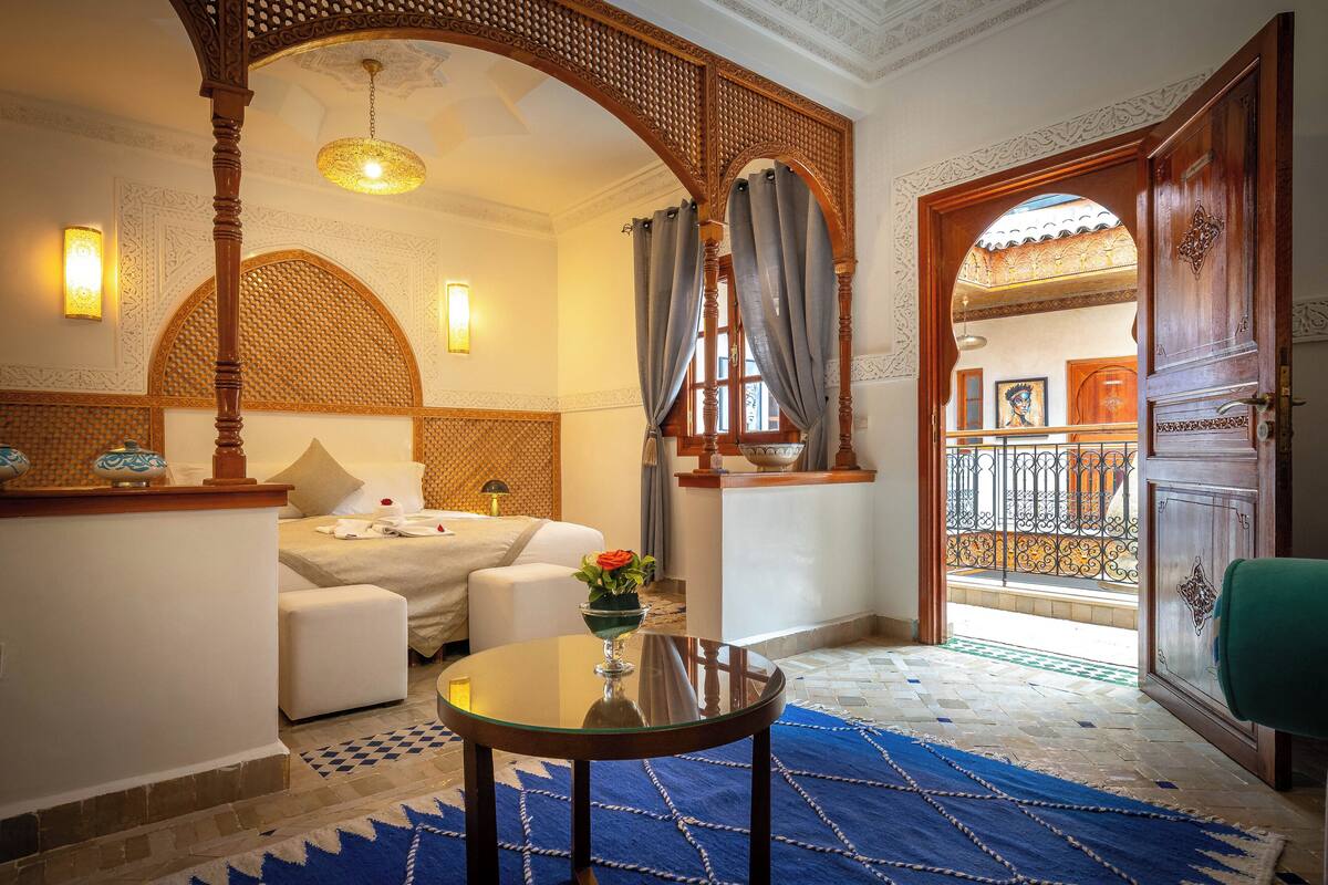 A room with a bed, a small table with a vase, a balcony with a railing, and a door with intricate woodwork.