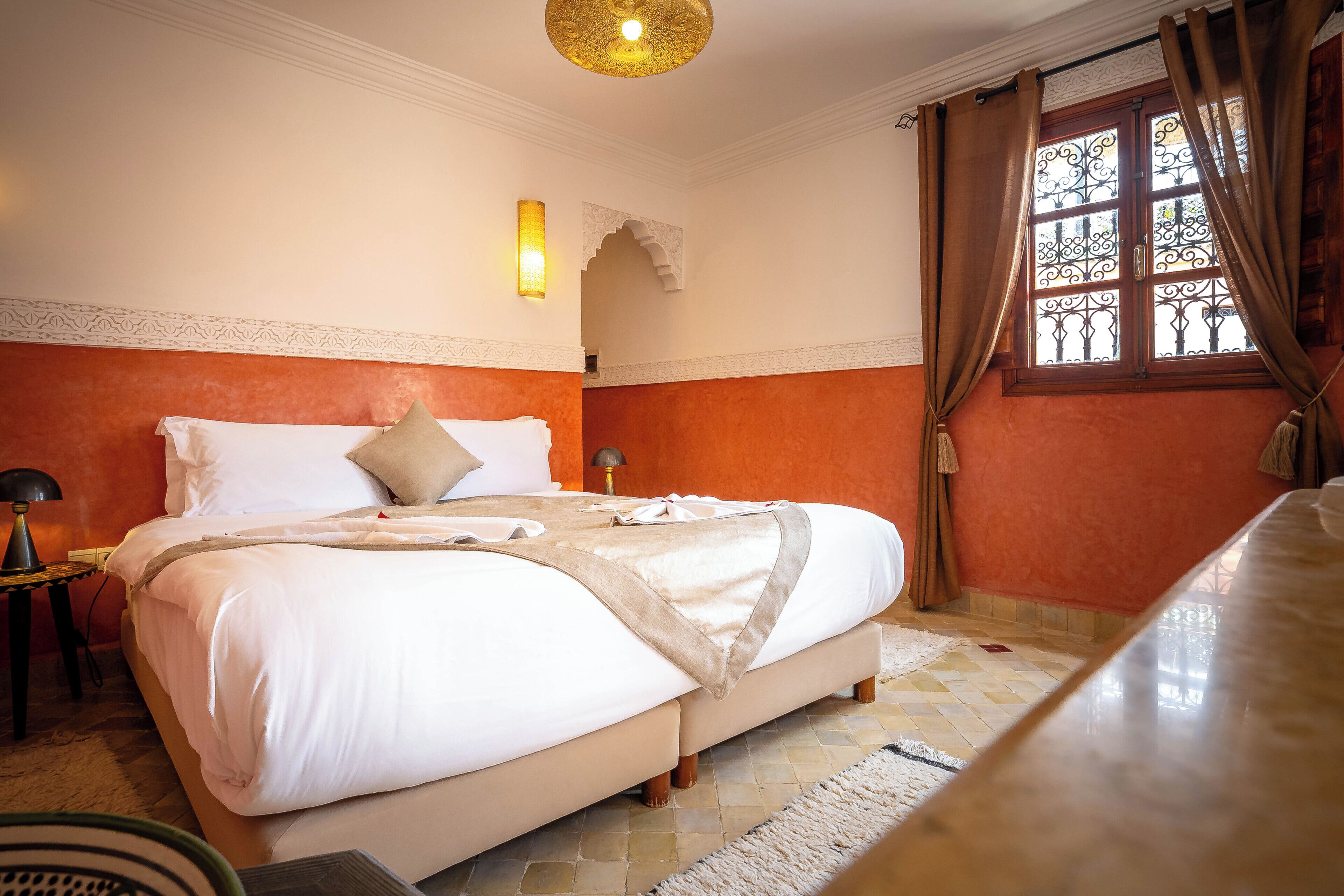 A neatly made bed with white linens and a beige throw, a bedside lamp, and a window with decorative iron bars and curtains.