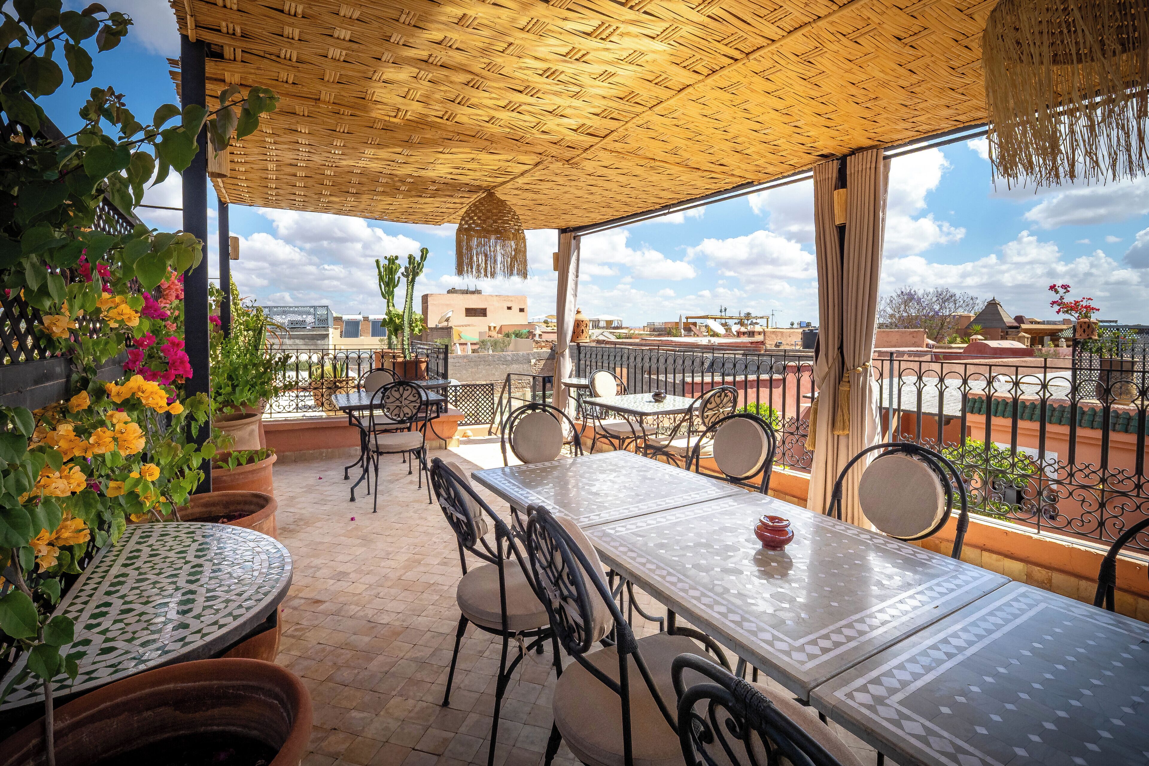 Un patio exterior con techo tejido, una mesa con sillas, plantas en macetas y vistas de la ciudad.