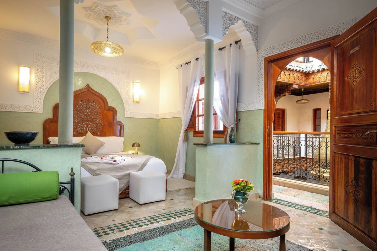 A room with a bed, a sitting area, a table with a vase, and a view of a balcony.