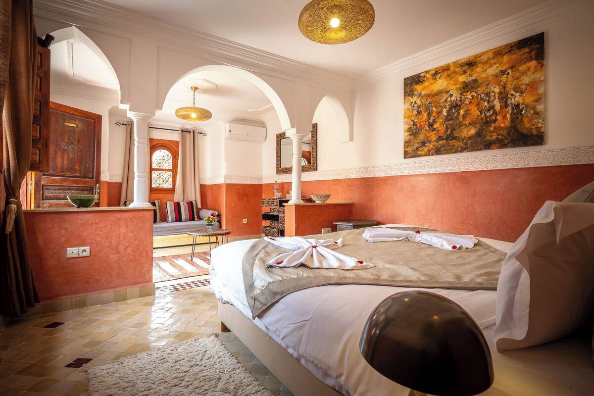 A hotel room with a large bed, a desk, a chair, a painting, and a view through an arched doorway.