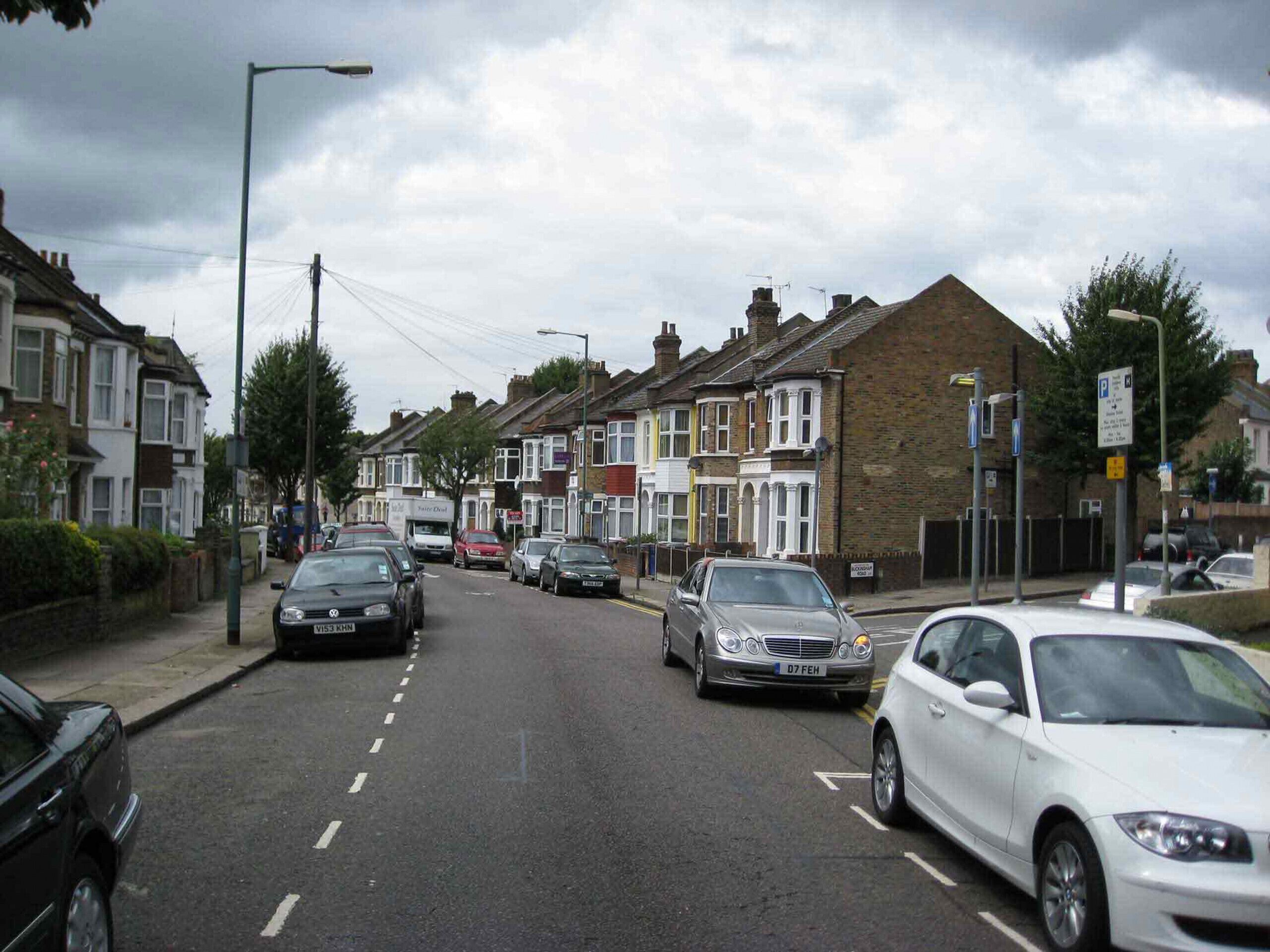 Une rue résidentielle bordée de maisons mitoyennes, avec des voitures garées et un ciel dégagé.