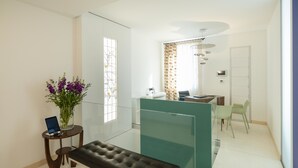 Una habitación de hotel moderna con un separador de vidrio, un banco, una pequeña mesa con una computadora portátil, un florero con flores, un baño con lavamanos y espejo, y una puerta.
