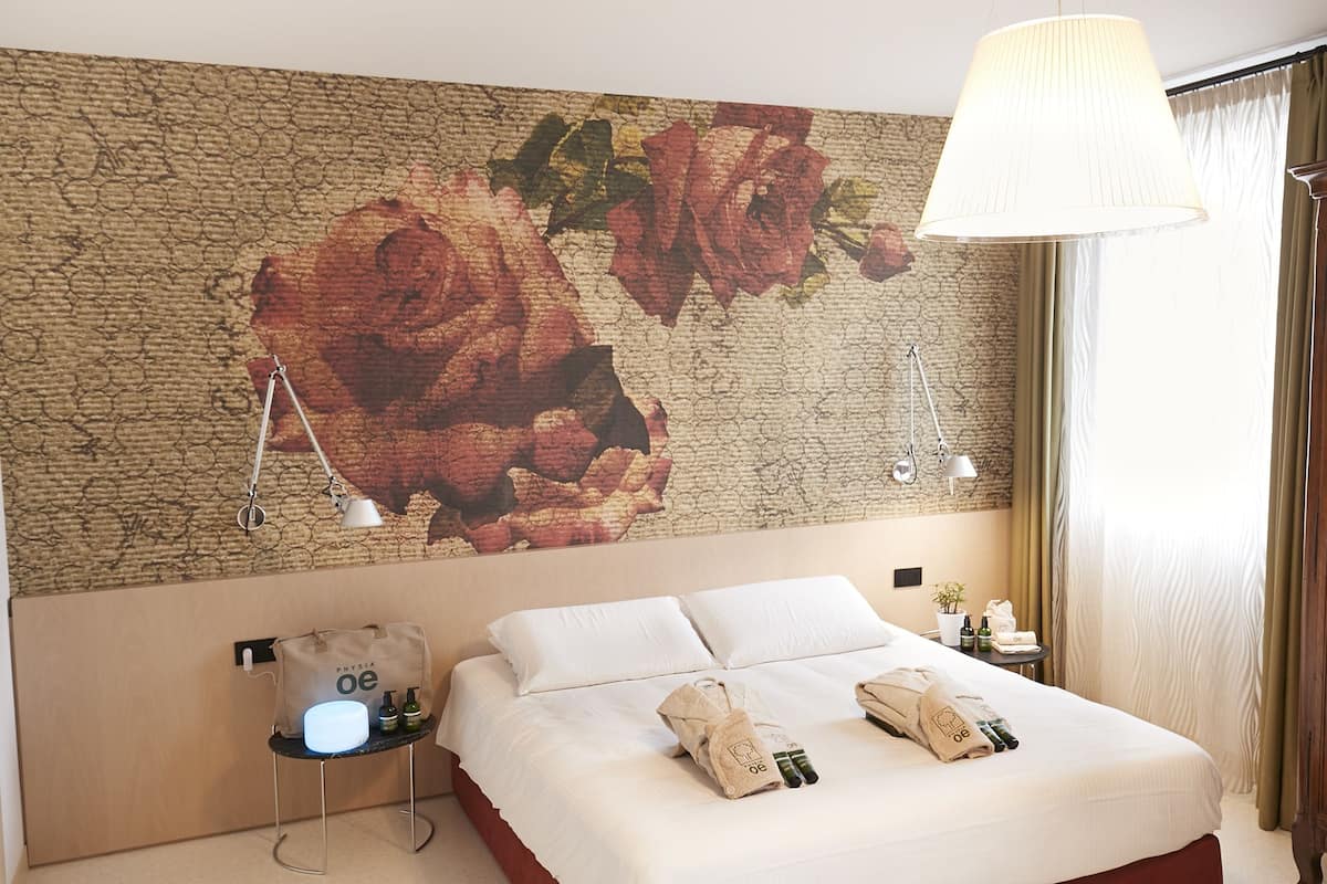 A hotel room with a large floral mural on the wall, a bed with white linens, a bedside table with a lamp, and a hanging light fixture.