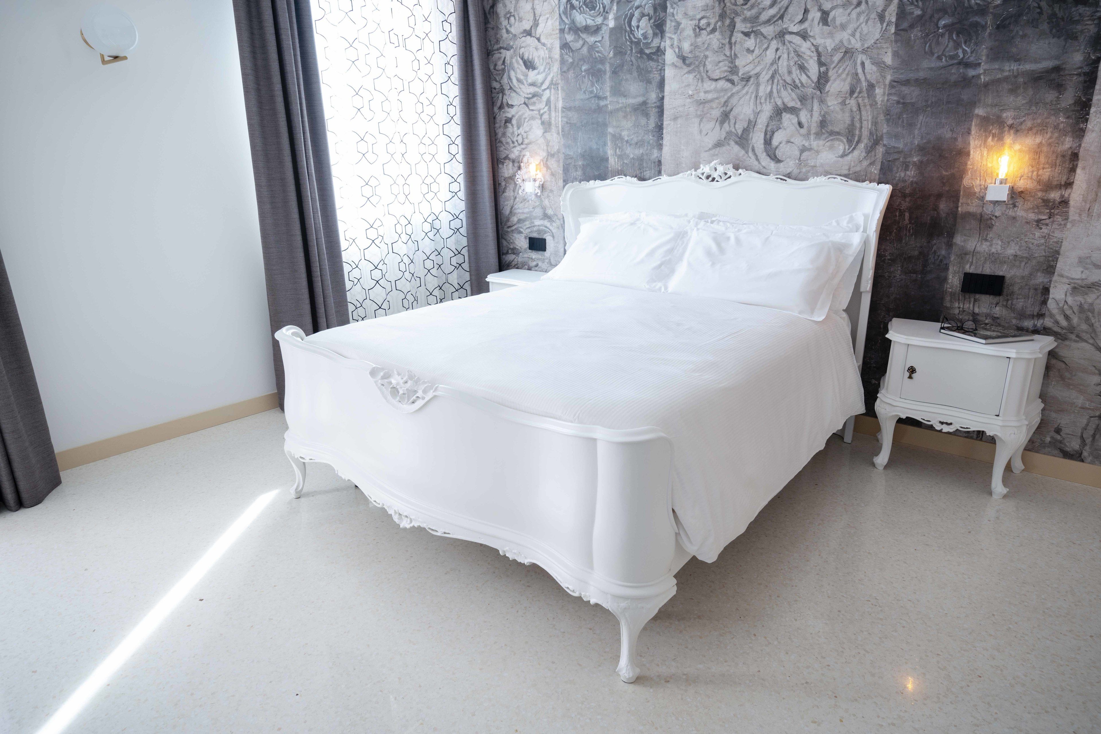 Una cama blanca en una habitación con pared de diseño marmolado y una mesita de noche con lámpara.