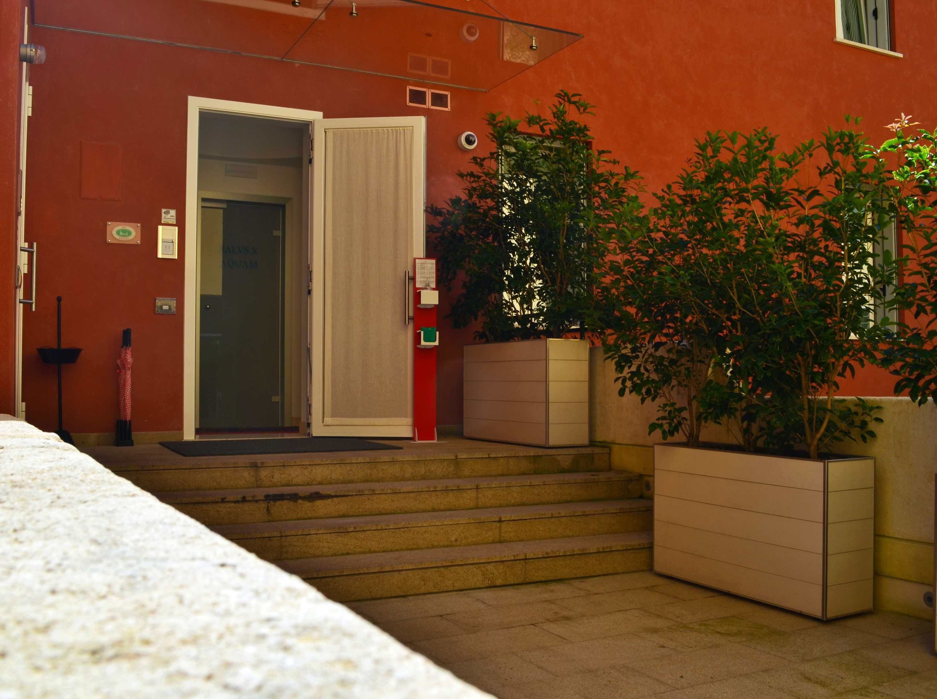 L’entrée d’un hôtel, avec une façade rouge, une porte en verre, des marches menant à l’entrée, un pot de fleurs et un extincteur.