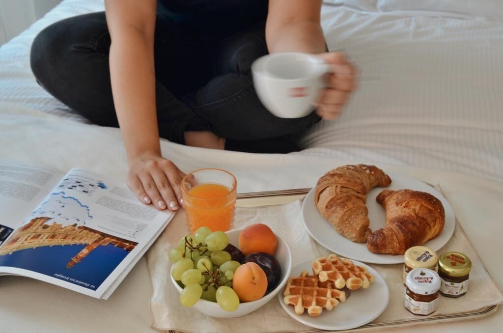 Uma pessoa sentada na cama com um livro, uma bandeja com café da manhã incluindo croissants, waffles, frutas e suco.
