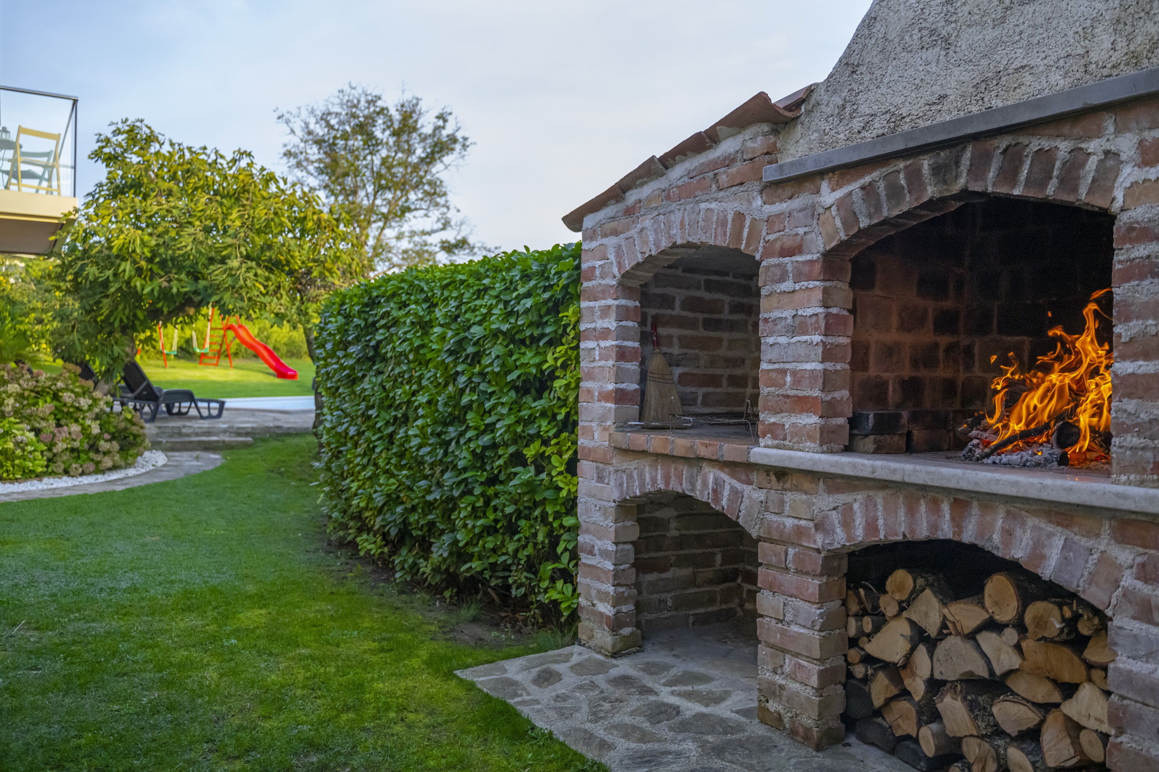 A brick outdoor kitchen with a fire, surrounded by a well-maintained garden and a playground.
