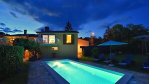 A swimming pool with a stone border, a small house with a lit window, and a lawn with lounge chairs.