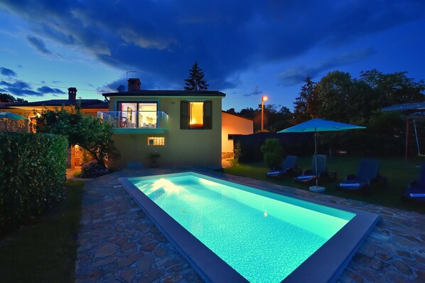A swimming pool with a stone border, a small house with a lit window, and a lawn with lounge chairs.