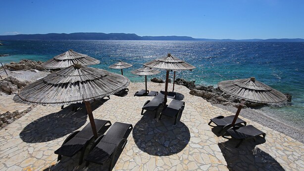 A beachside area with stone flooring, several thatched umbrellas, and lounge chairs.