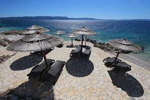 A beachside area with stone flooring, several thatched umbrellas, and lounge chairs.