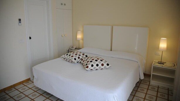 A neatly made bed with white bedding and polka-dotted pillows, a white headboard, and bedside lamps on a tiled floor. - Via Capo Rooms (Sorrento)