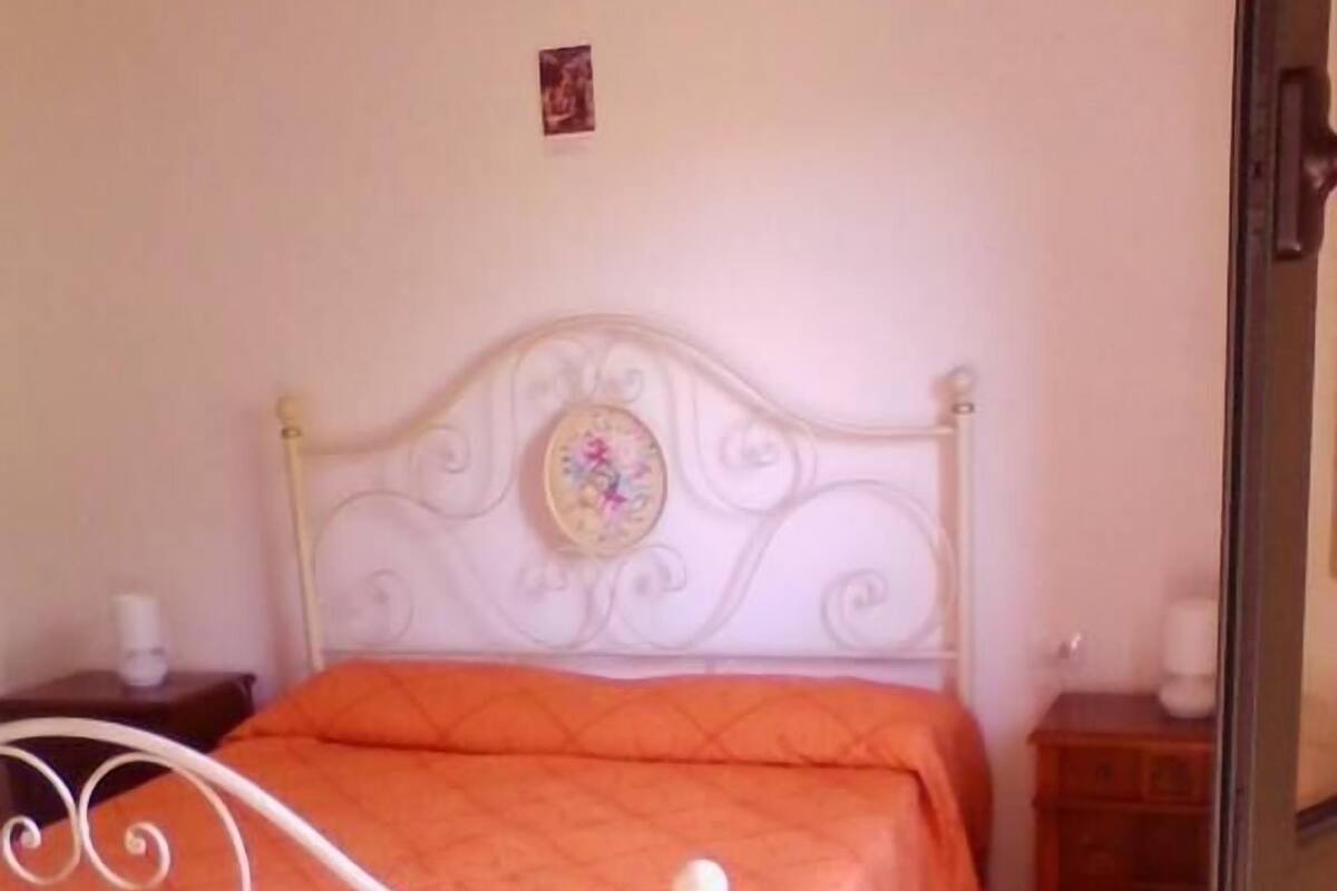 A bedroom with a white metal bed frame, an orange bedspread, a small bedside table with a lamp, and a wall-mounted picture.