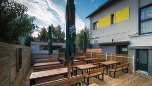 Un patio al aire libre con bancos de madera y sombrillas verdes, una cerca de madera y un edificio con una franja amarilla.