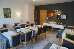 A dining area with multiple tables and chairs, a buffet setup, and a wall-mounted shelf with glassware. - Gästehaus Kiebel (Lösnich)