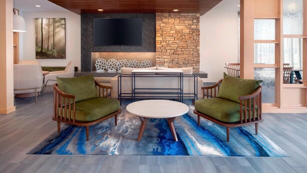 A modern living room with a round table, two green armchairs, a stone fireplace, and a large TV mounted on the wall. - Fairfield Inn & Suites by Marriott Denver Tech Center North (Denver)