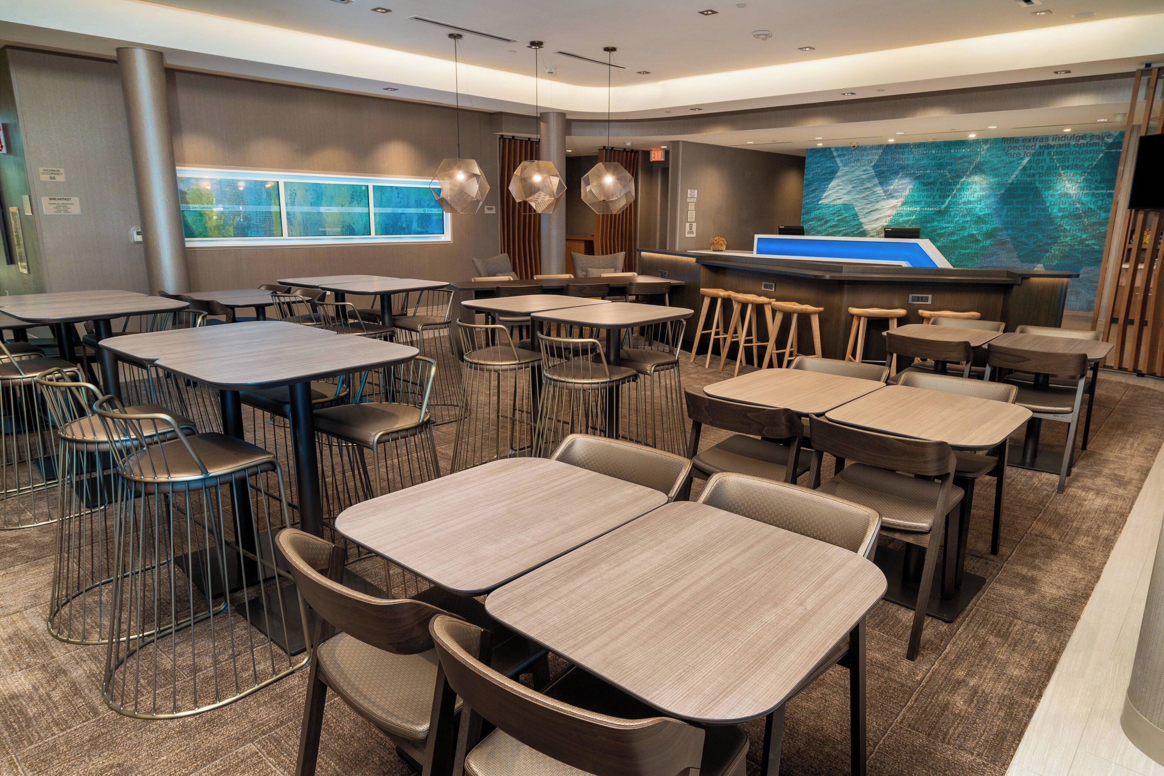 A modern hotel lobby with multiple tables and chairs, a reception desk, and a large digital screen displaying a map.
