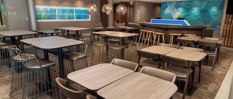 A modern hotel lobby with multiple tables and chairs, a reception desk, and a large digital screen displaying a map.