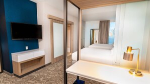 Et hotelværelse med et vægmonteret fladskærms-TV, et hvidt natbord med lampe, en seng og havudsigt gennem et vindue.