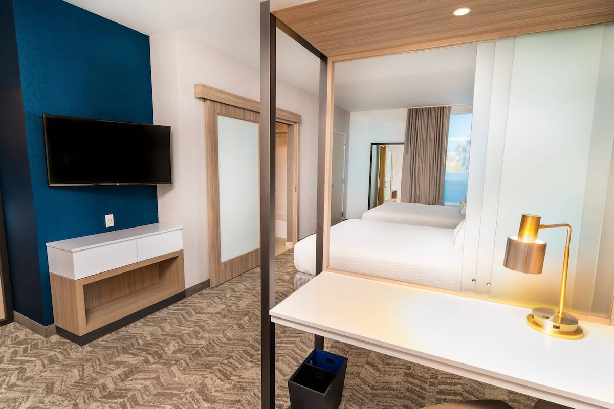 Hotellihuone, jossa on seinälle asennettu taulutelevisio, valkoinen yöpöytä lampun kanssa, sänky ja ikkunasta avautuva näkymä merelle.