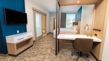 A hotel room with a large bed, a desk with a chair, a flat-screen TV mounted on the wall, and a view of the outdoors through the window.
