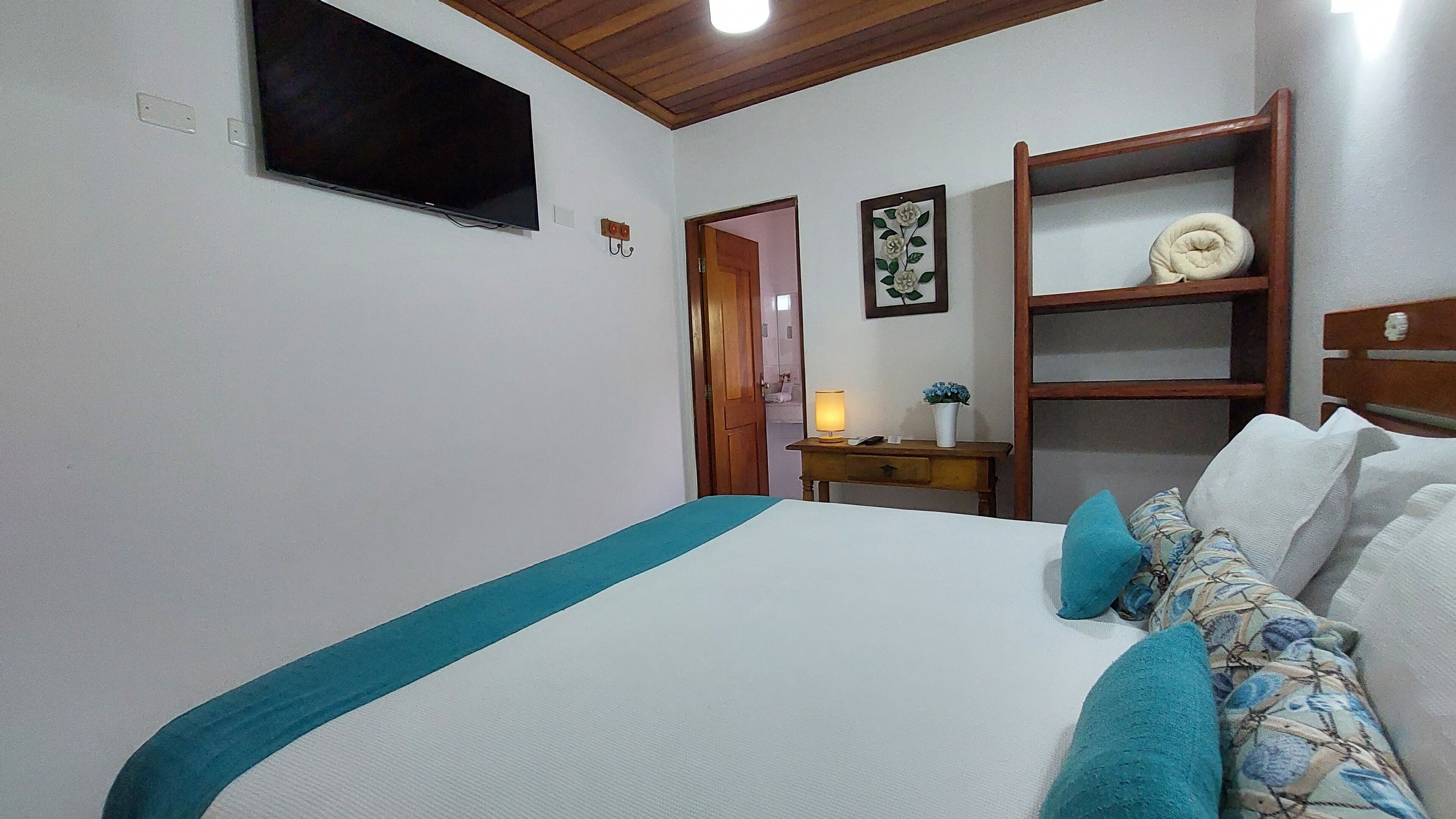 Un dormitorio con cama, una mesita de noche con lámpara, un televisor de pared y un estante de madera con un objeto decorativo.