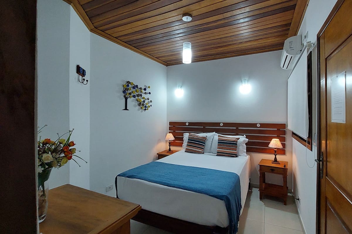 Une chambre avec un plafond en bois, un lit recouvert d’une courtepointe bleue, deux tables de chevet avec des lampes et une armoire en bois.