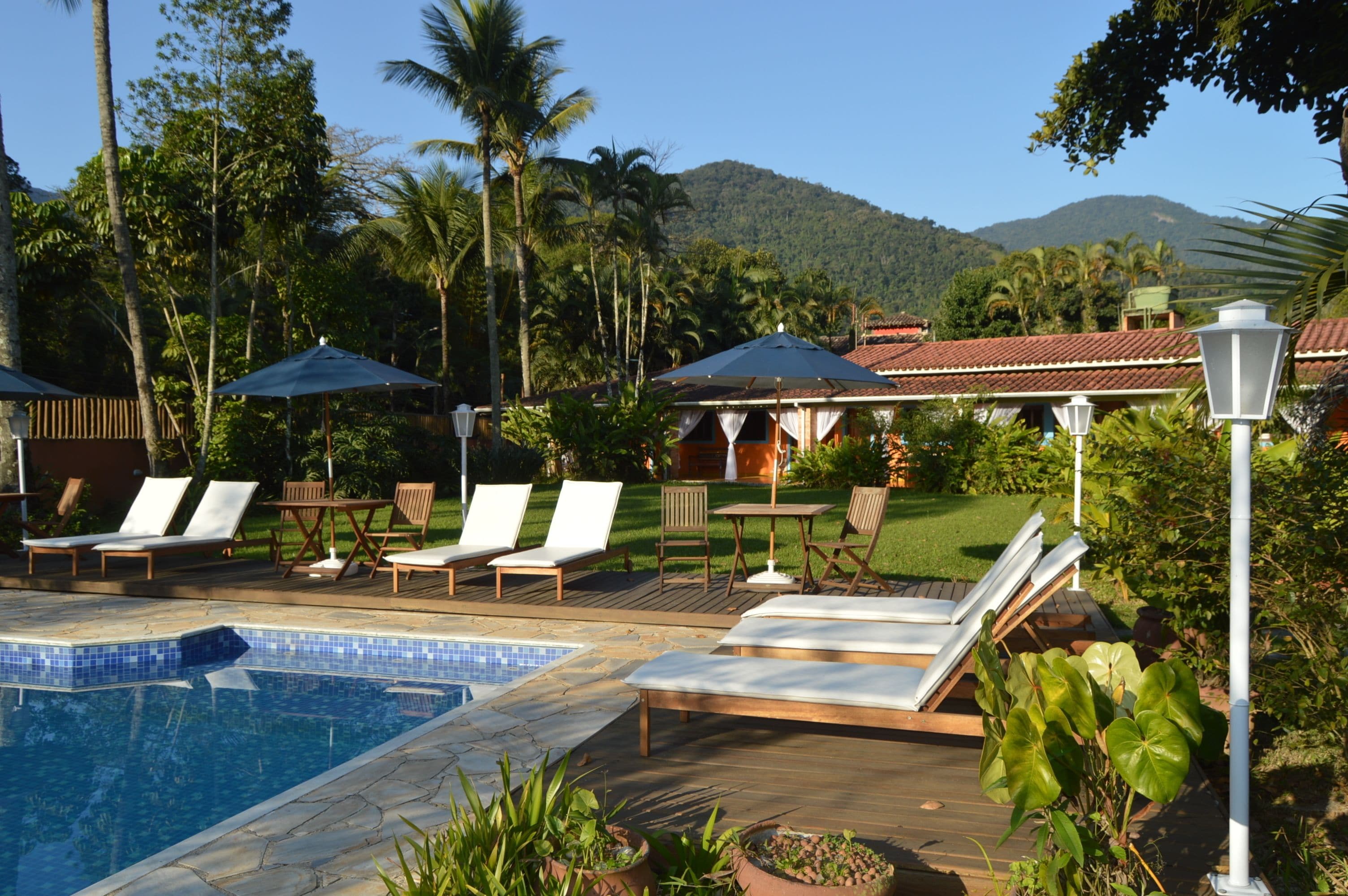 Uma área de piscina com espreguiçadeiras e guarda-sóis, cercada por vegetação exuberante e uma montanha ao fundo.