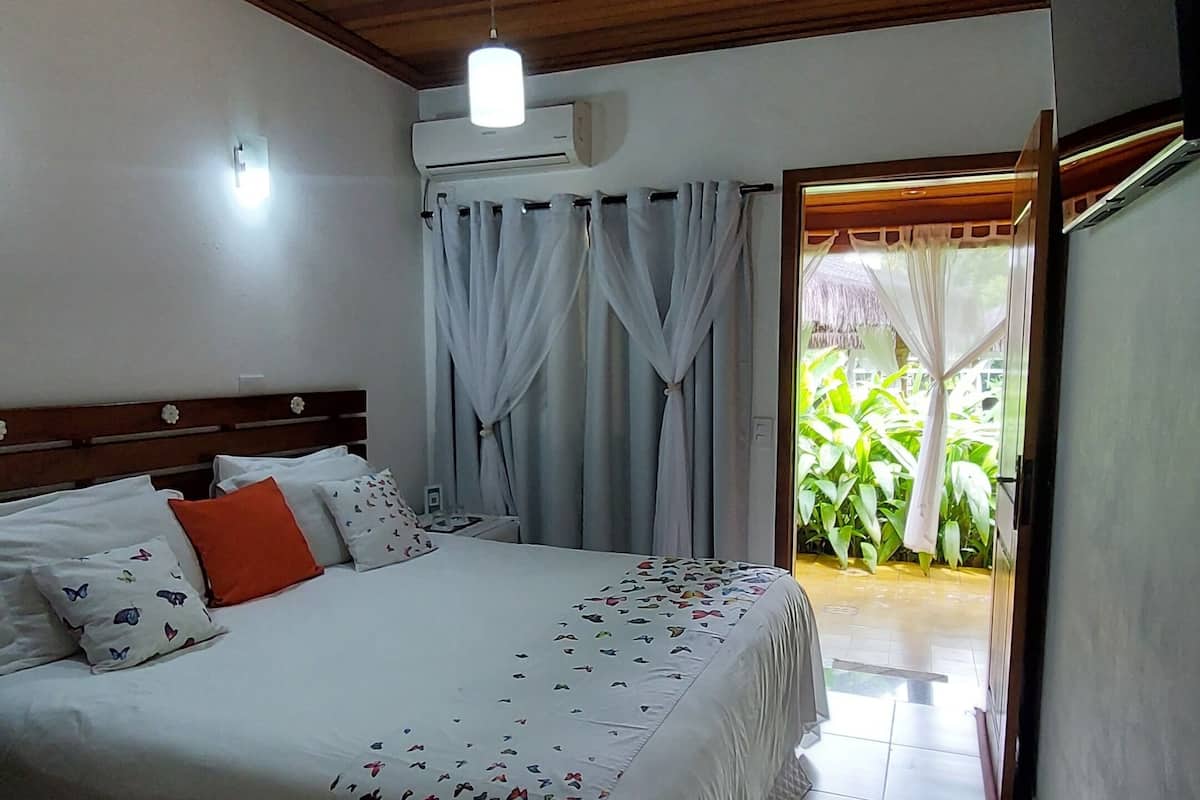 Une chambre avec un plafond en bois, un lit recouvert d’une literie à motifs, une table de chevet et une vue sur un jardin par une porte ouverte.
