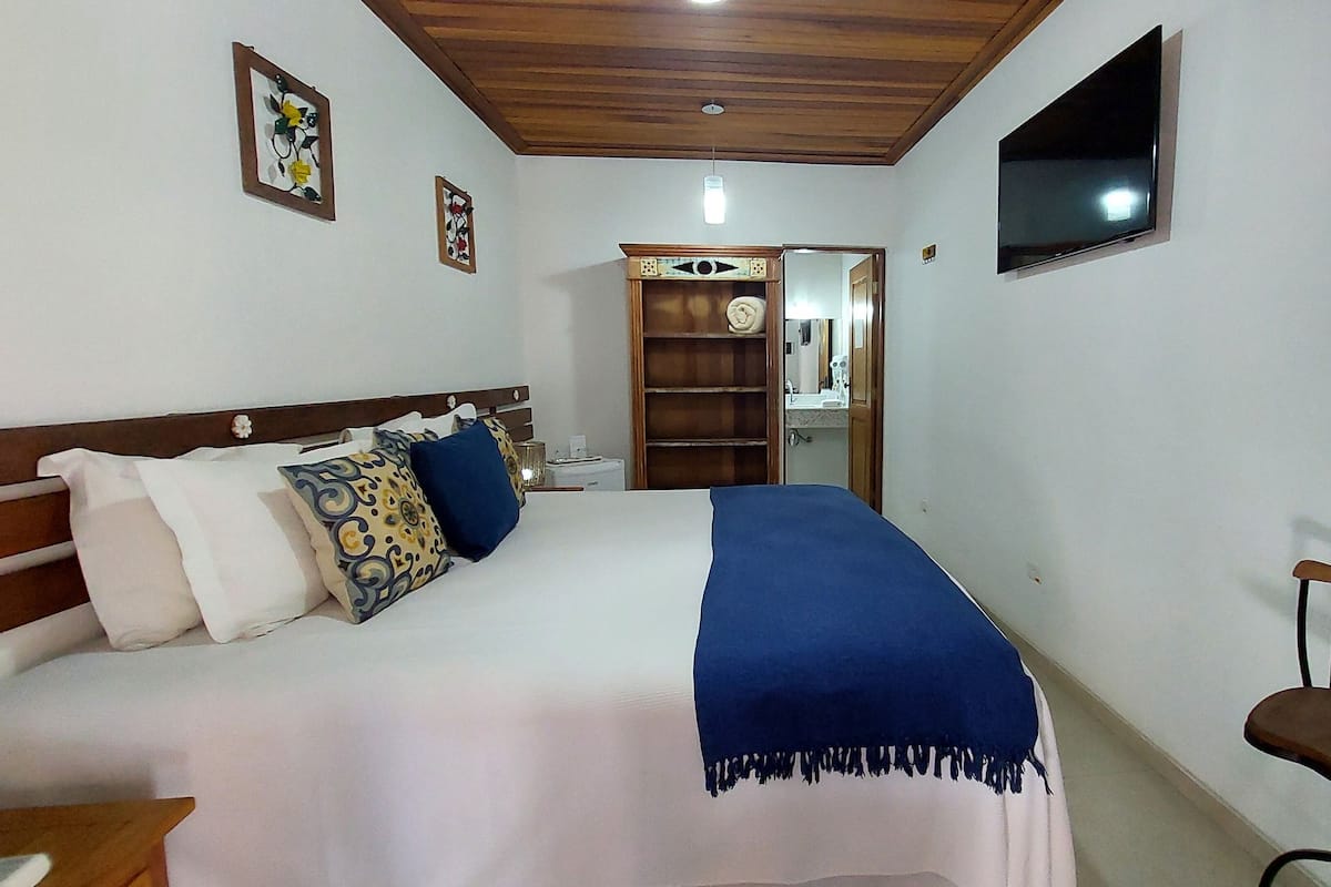 Une chambre à coucher comprenant un lit, un bureau, une chaise, un téléviseur et une salle de bain visible à travers une porte ouverte.