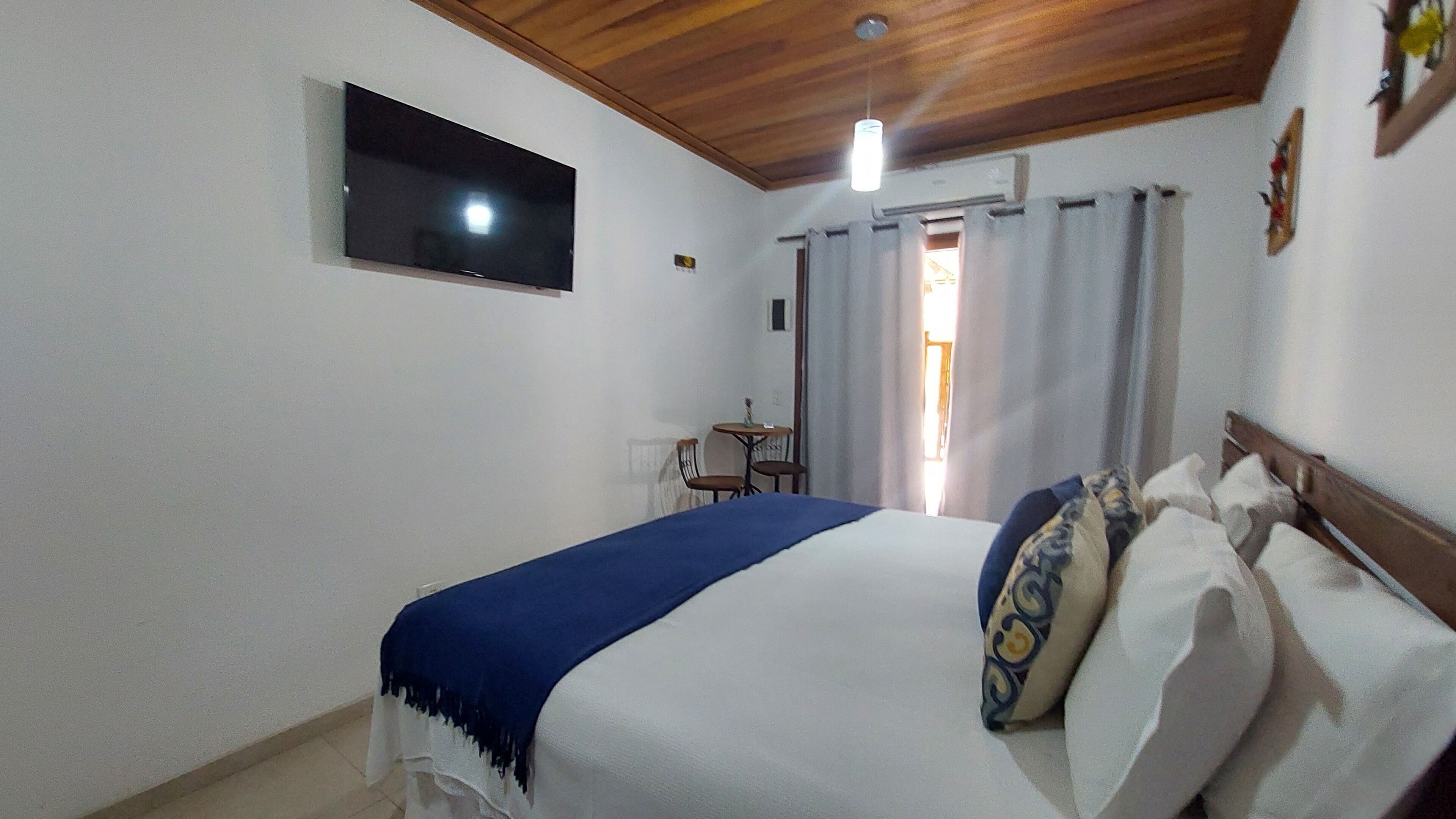 A bedroom with a bed, a TV mounted on the wall, a small table with chairs, and a window with curtains.
