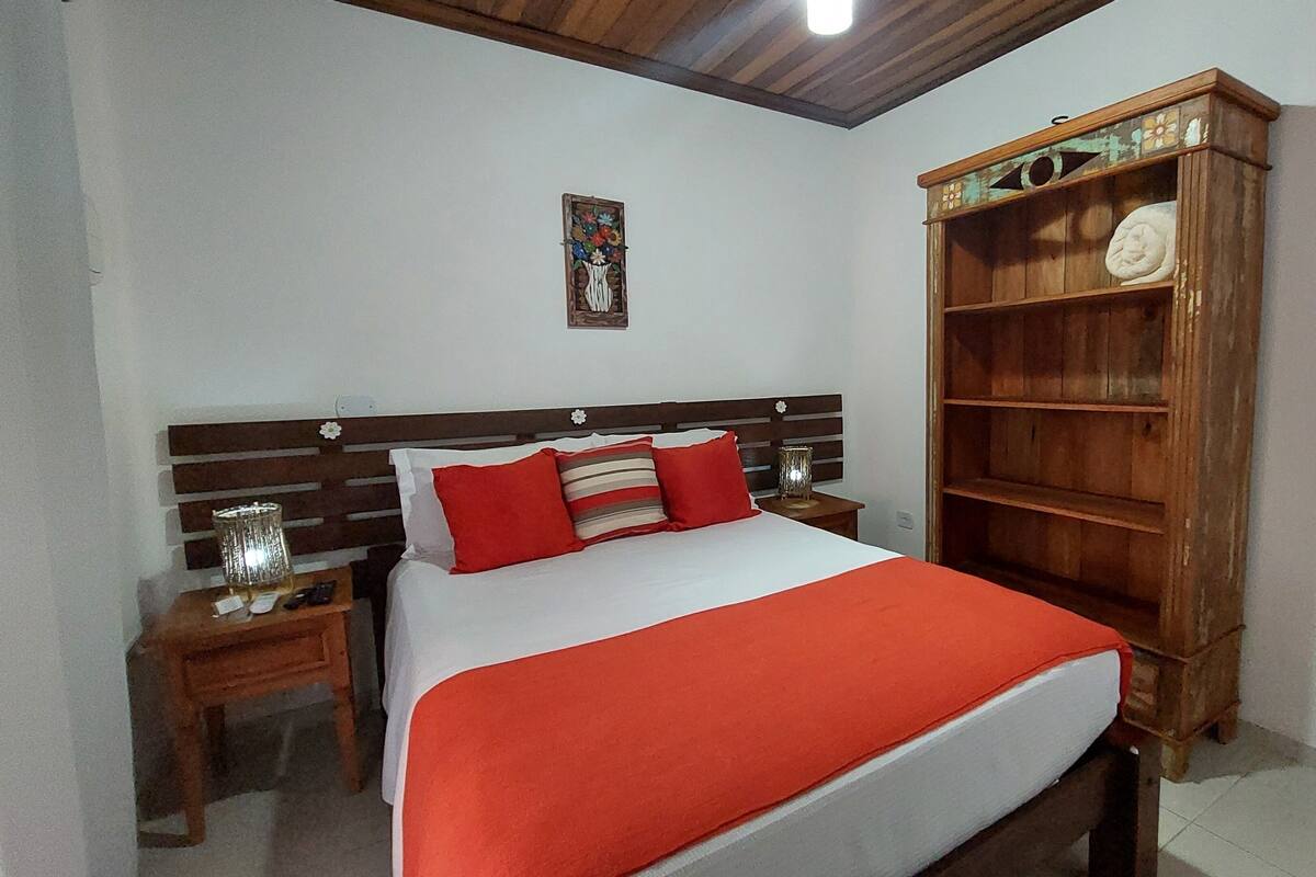 Une chambre à coucher avec un lit, une tête de lit en bois, des tables de chevet, une couverture rouge, un élément décoratif mural et une bibliothèque en bois.