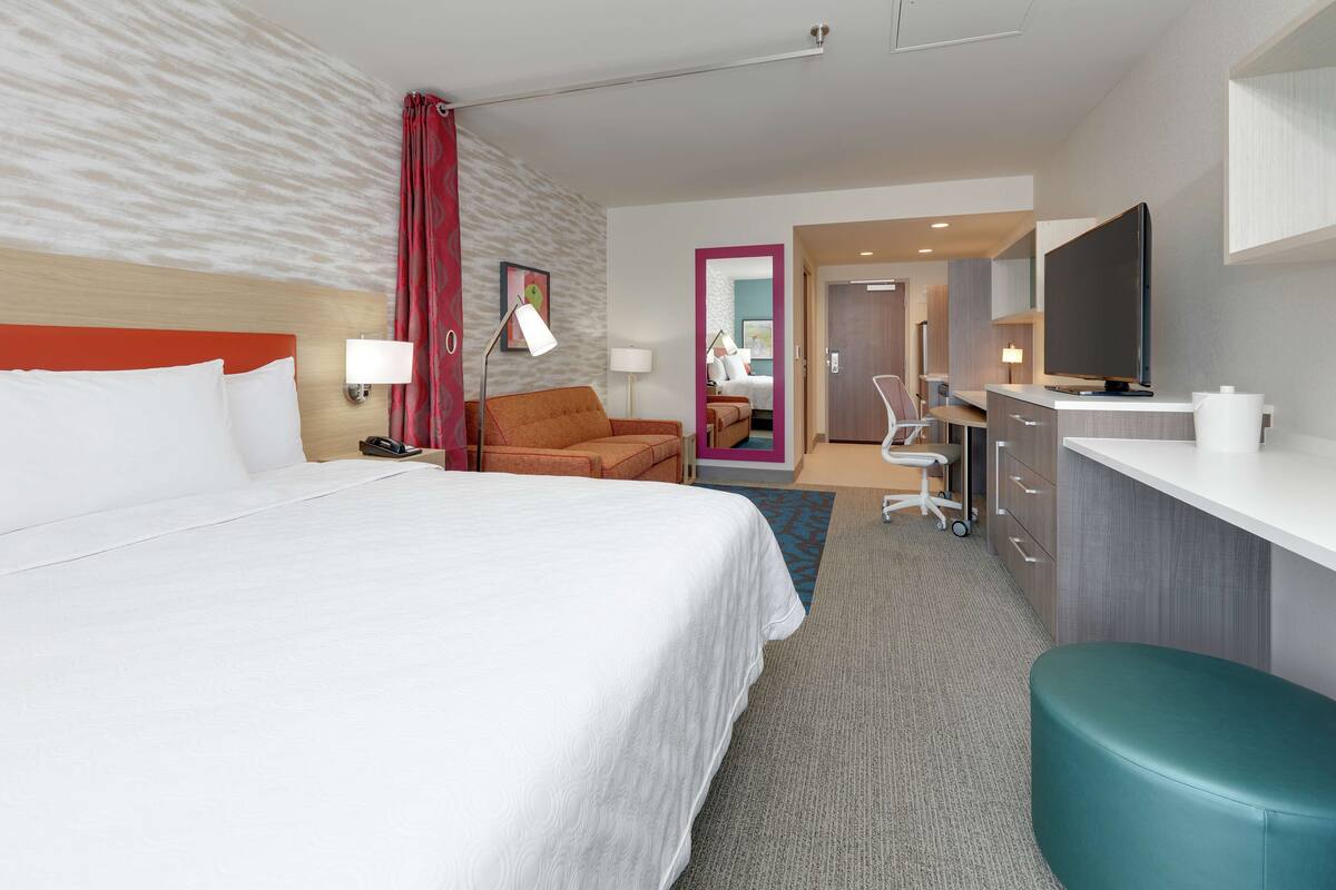 Une chambre d’hôtel avec un grand lit, un bureau doté d’un téléviseur à écran plat, une chaise, une lampe de bureau et une salle de bain visible à travers une porte ouverte.