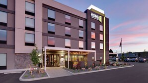 A modern hotel building with a prominent 'HOME2' sign, a well-lit entrance, and a landscaped area with plants and seating.