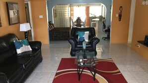 A living room with a black leather sofa, a glass coffee table, and a red and beige patterned rug. There is a black leather armchair with a blue pillow, and a dining area in the background. - Villa Edinburgh (Montego Bay)