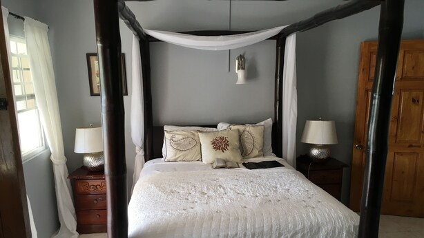 A four-poster bed with a white bedspread and decorative pillows, flanked by two bedside tables with lamps, in a room with light blue walls and a wooden door. - Villa Edinburgh (Montego Bay)