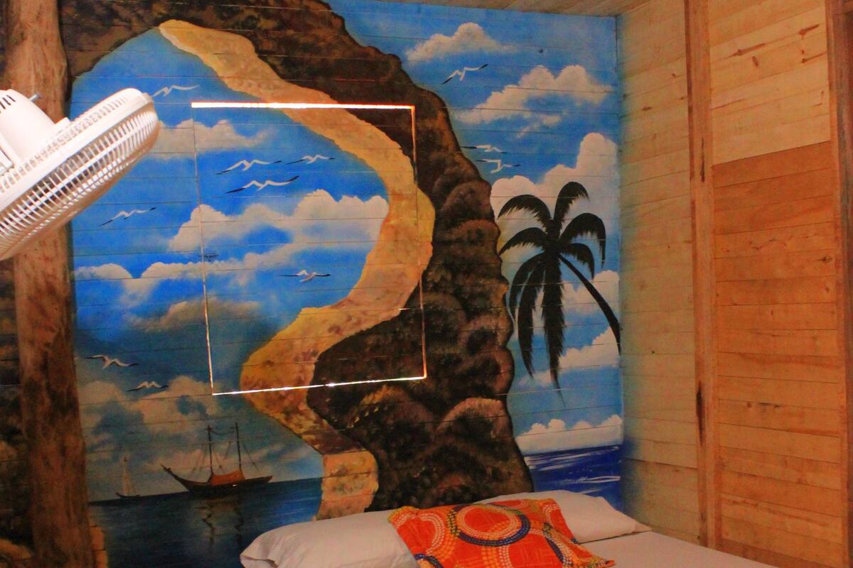 Une chambre avec un lit, un ventilateur et une fresque représentant une scène de plage.