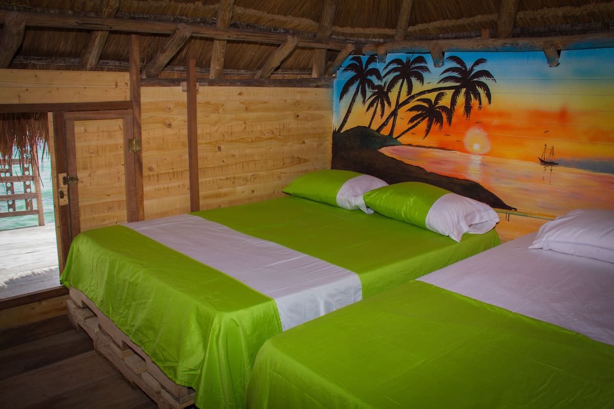 Une chambre avec deux lits, chacun agrémenté de draps verts et blancs, des murs en bois et une fresque représentant un coucher de soleil avec des palmiers.