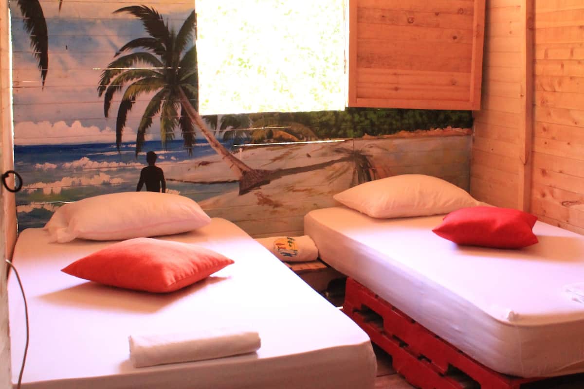 Une chambre avec deux lits, chacun équipé d’un oreiller et d’une couverture, ainsi qu’un ventilateur de plafond et une fresque murale représentant une scène de plage.