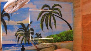 A room with two beds, each with a red pillow and white sheets. A large beach mural on the wall depicts palm trees and the ocean.