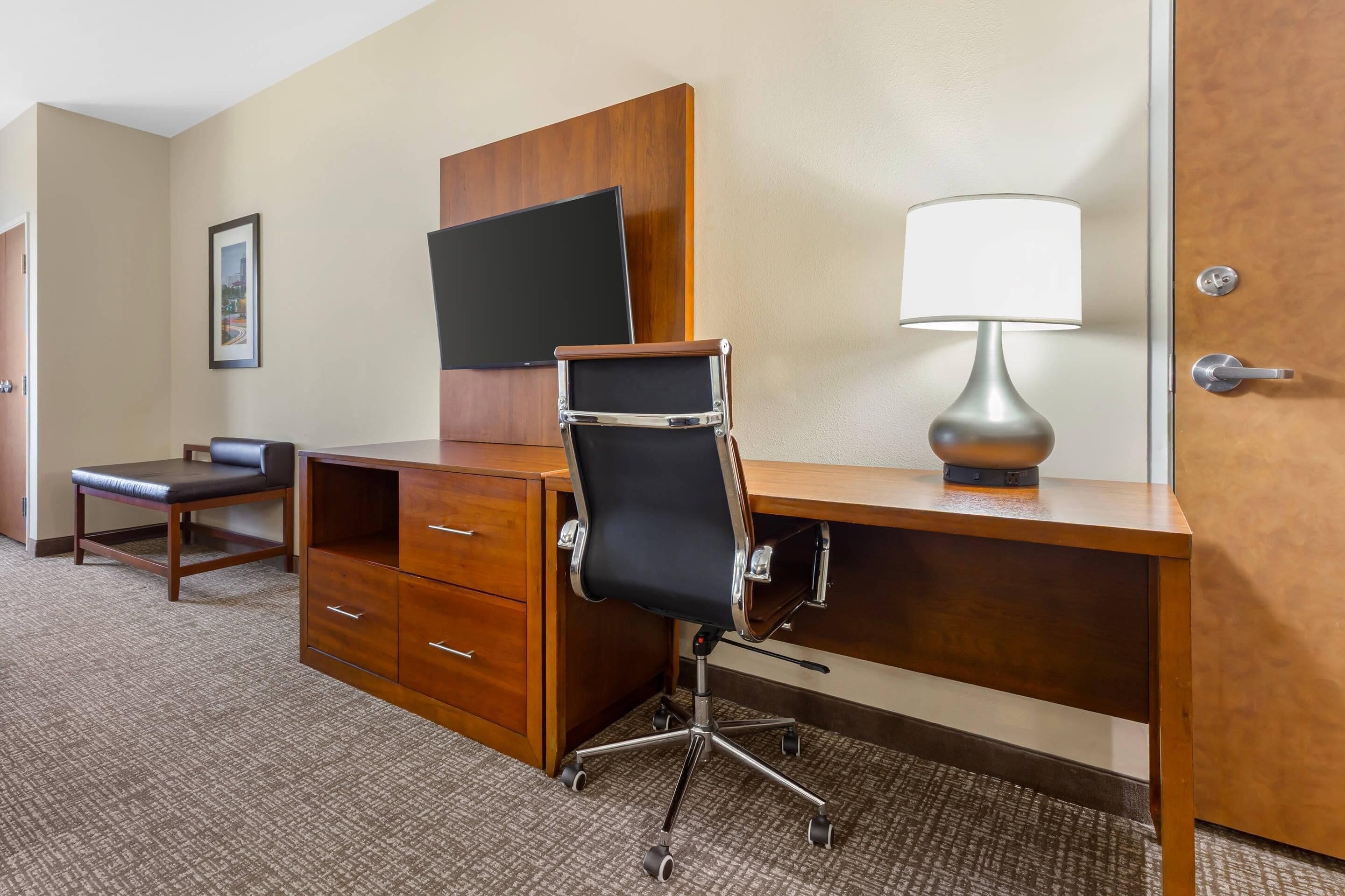 A hotel room with a wooden desk, a black office chair, a flat-screen TV mounted on the wall, and a table lamp.