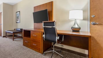 A hotel room with a wooden desk, a black office chair, a flat-screen TV mounted on the wall, and a table lamp.