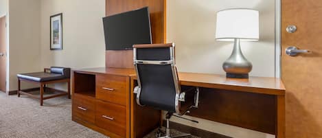 A hotel room with a wooden desk, a black office chair, a flat-screen TV mounted on the wall, and a table lamp.