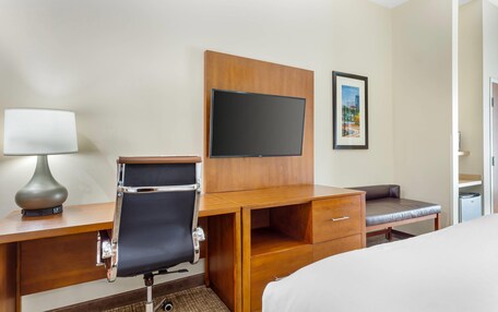 A hotel room with a wooden desk, a chair, a bed, a TV, and a framed picture on the wall.