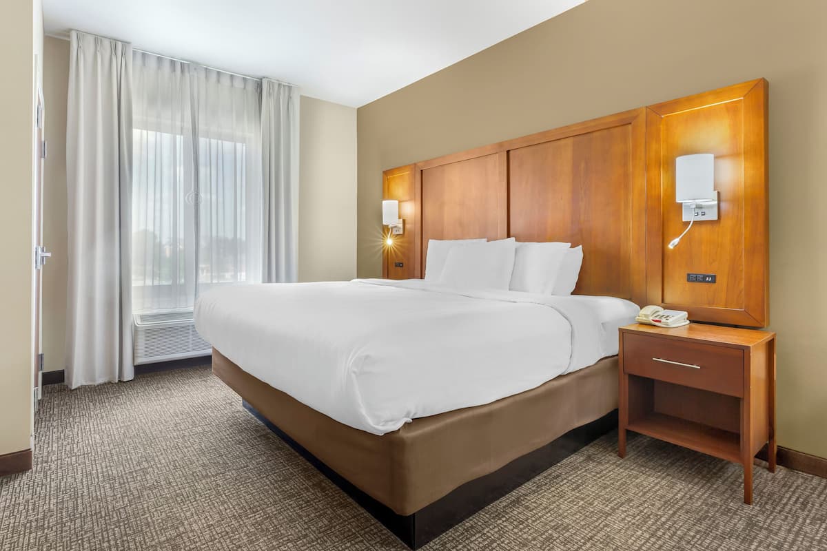 A hotel room with a large bed, a wooden headboard, a nightstand with a phone, and a window with sheer curtains.