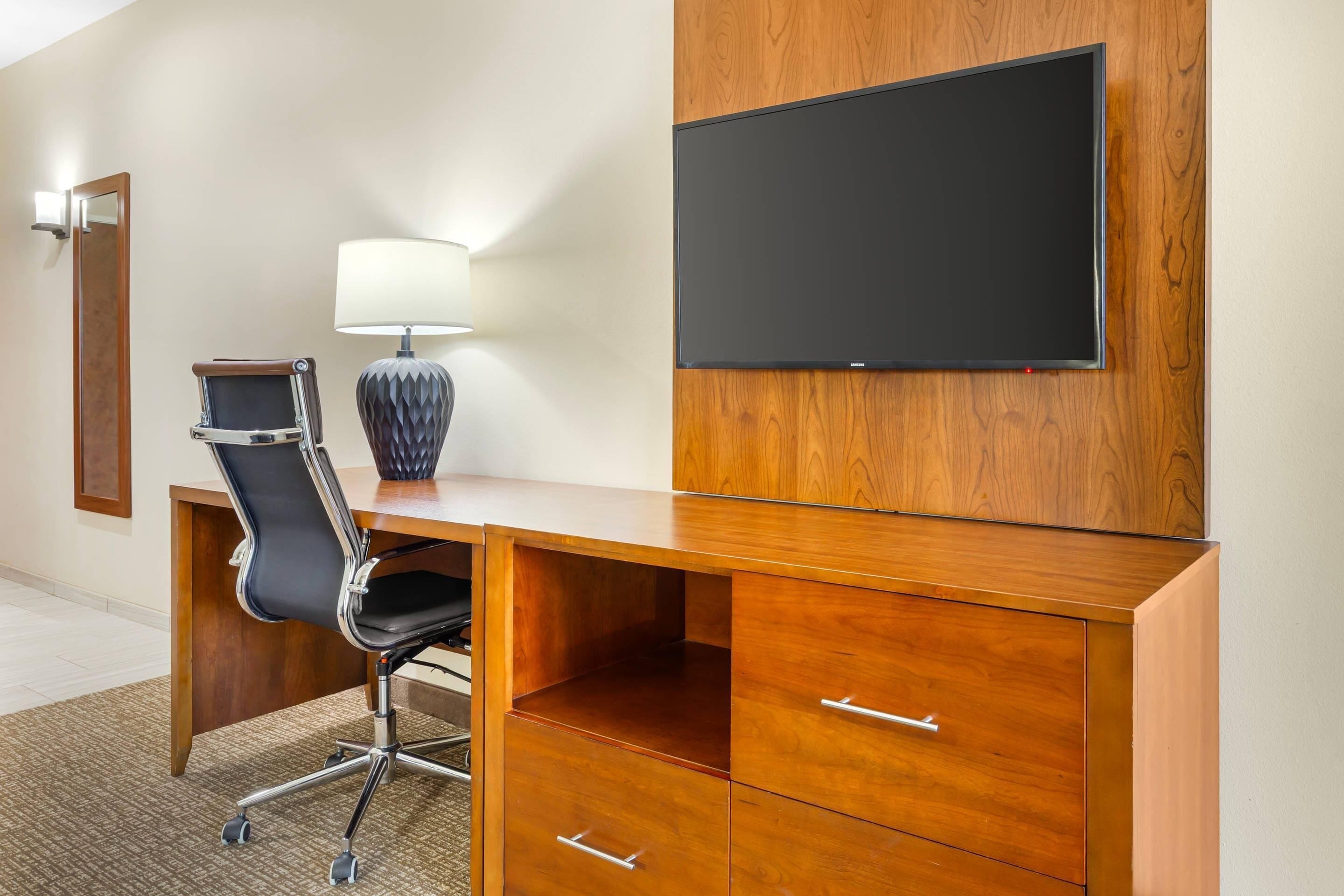 A hotel room with a wooden desk, a black office chair, a flat-screen TV mounted on the wall, a lamp, and a mirror.