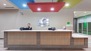 A reception desk with a logo for Holiday Inn, a woman standing behind it, and a modern ceiling design. - Holiday Inn Amarillo East (Amarillo)