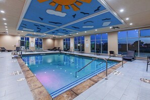An indoor swimming pool with clear water, surrounded by lounge chairs and a ceiling with a sun and cloud design.